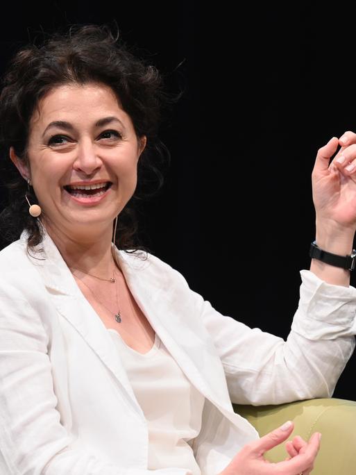 Ece Temelkuran spricht lächelnd auf einem Podium und trägt weiße Kleidung vor dunklem Hintergrund