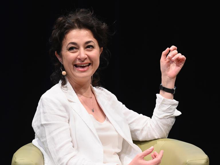 Ece Temelkuran spricht lächelnd auf einem Podium und trägt weiße Kleidung vor dunklem Hintergrund