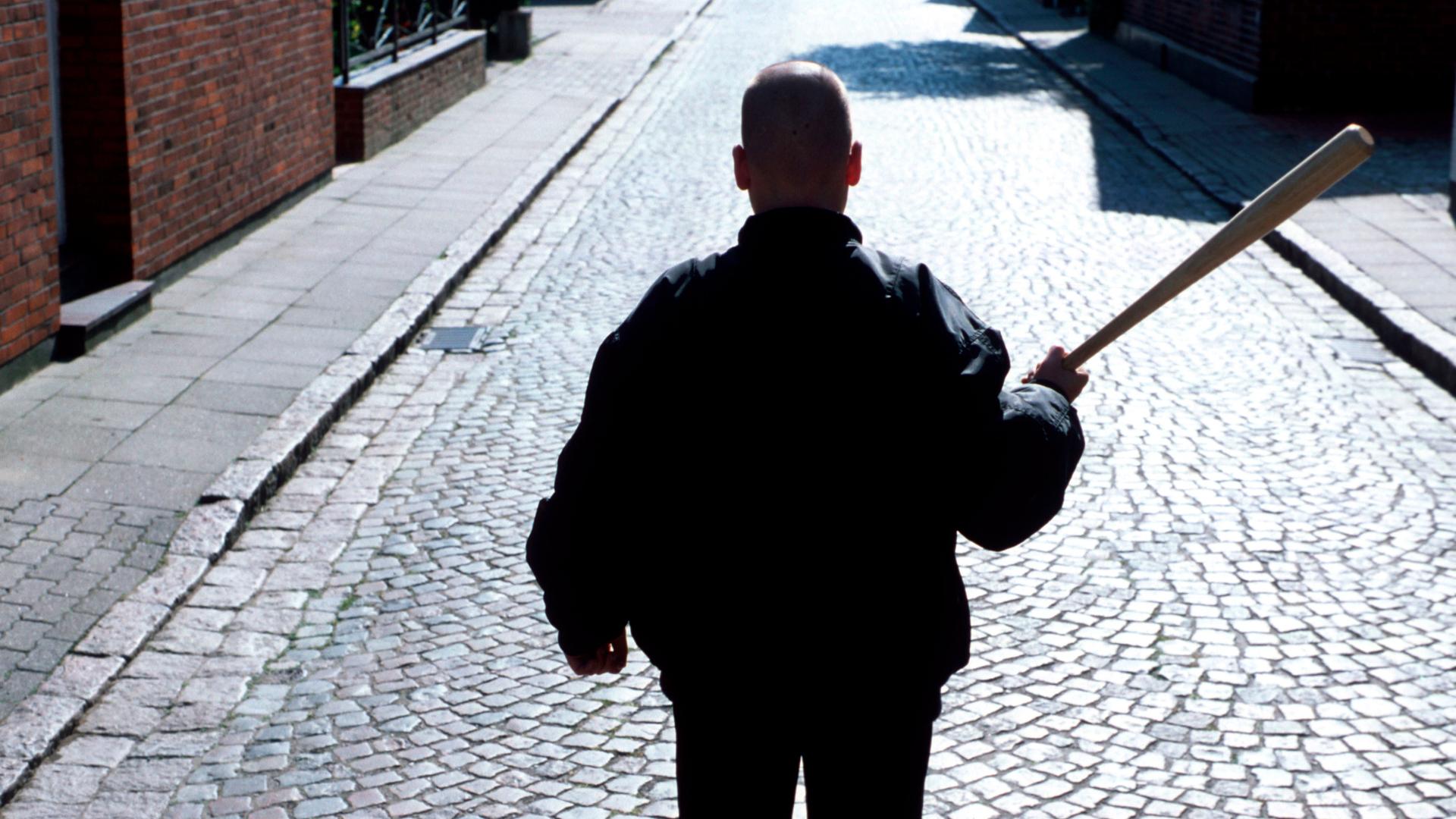 Ein Neonazi mit Baseballschlaeger und Bomberjacke steht mit dem Rücken zur Kamera auf einer Straße mit Kopfsteinpflaster.