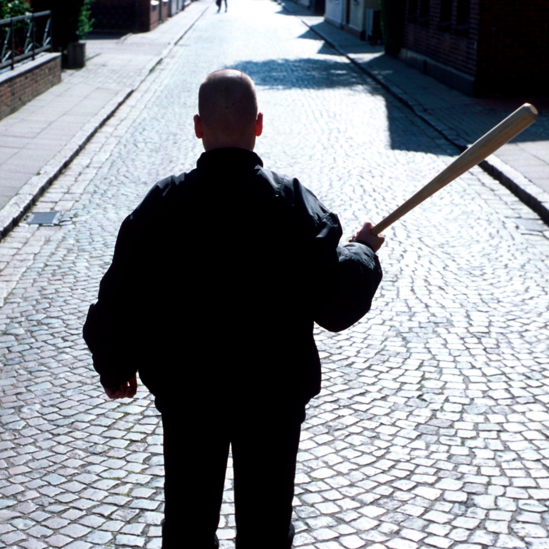 Ein Neonazi mit Baseballschlaeger und Bomberjacke steht mit dem Rücken zur Kamera auf einer Straße mit Kopfsteinpflaster.