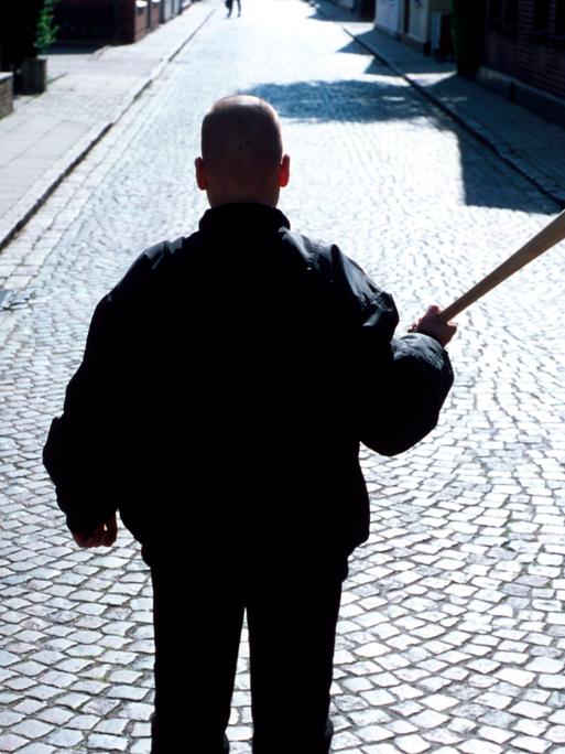 Ein Neonazi mit Baseballschlaeger und Bomberjacke steht mit dem Rücken zur Kamera auf einer Straße mit Kopfsteinpflaster.
