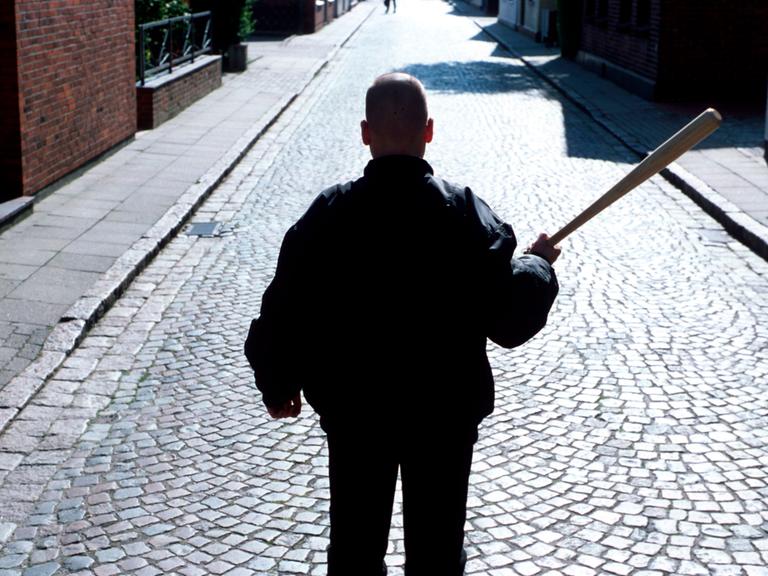 Ein Neonazi mit Baseballschlaeger und Bomberjacke steht mit dem Rücken zur Kamera auf einer Straße mit Kopfsteinpflaster.