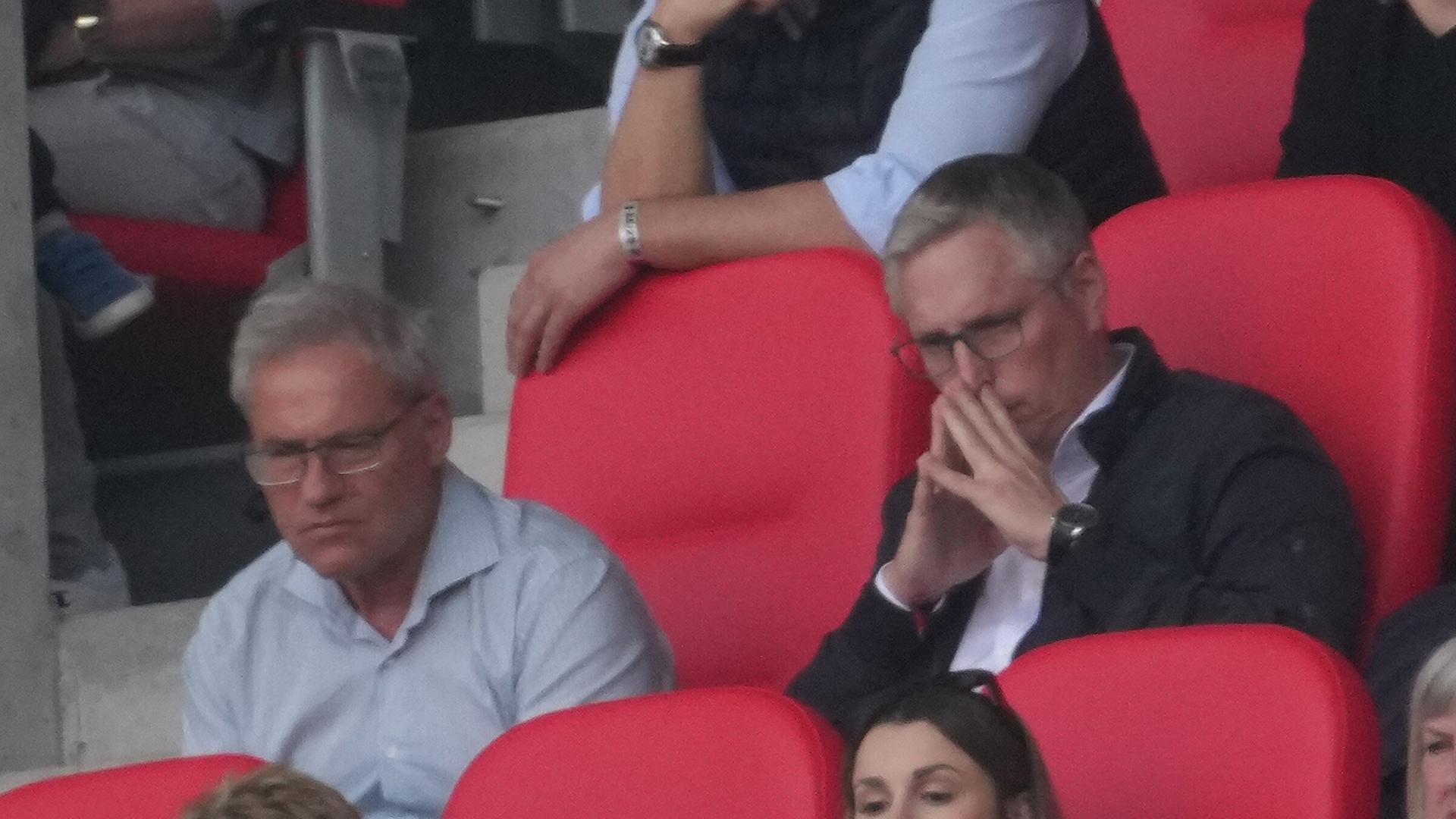 Die beiden sitzen nebeneinander auf einer Stadiontribüne in roten komfortablen Sitzen. 