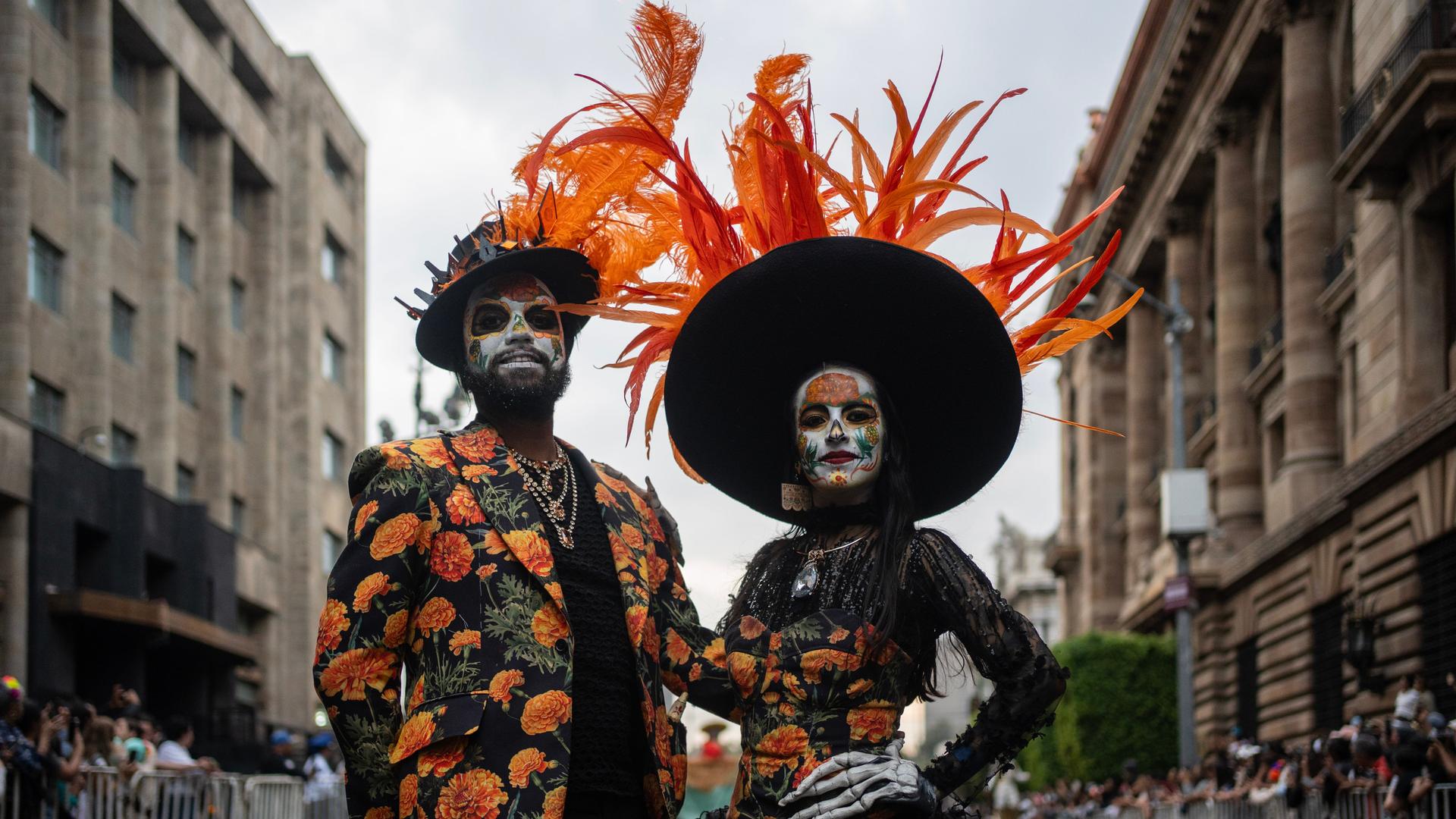 Ein Mann und eine Frau haben sich für den Tag der Toten verkleidet. Ihre Gesichter sind weiß geschminkt. Sie tragen bunte Kleidung. Die beiden schauen in Richtung der Kamera.  Ein Mann und eine Frau haben sich für den Tag der Toten verkleidet. Ihre Gesichter sind weiß geschminkt. Sie tragen bunte Kleidung. Die beiden schauen in Richtung der Kamera.