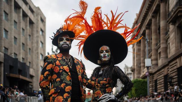 Ein Mann und eine Frau haben sich für den Tag der Toten verkleidet. Ihre Gesichter sind weiß geschminkt. Sie tragen bunte Kleidung. Die beiden schauen in Richtung der Kamera. 