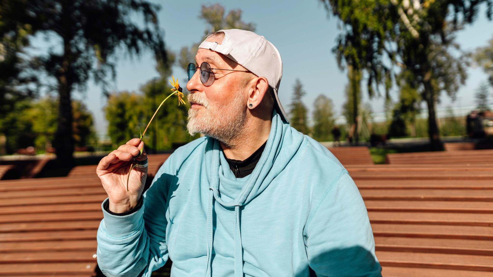 Ein älterer Mann sitzt im Sommer auf einer Parkbank und riecht an einer Blume. Ein älterer Mann sitzt im Sommer auf einer Parkbank und riecht an einer Blume.