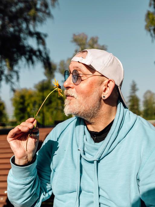 Ein älterer Mann sitzt im Sommer auf einer Parkbank und riecht an einer Blume.