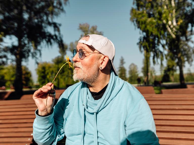 Ein älterer Mann sitzt im Sommer auf einer Parkbank und riecht an einer Blume.