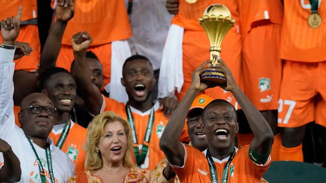 Mehrere Spieler von der Elfenbein-Küste stehen nebeneinander. Sie tragen orange-farbene Trikots. Ein Spieler hält den goldenen Pokal in den Händen. Neben ihm steht eine Frau.
