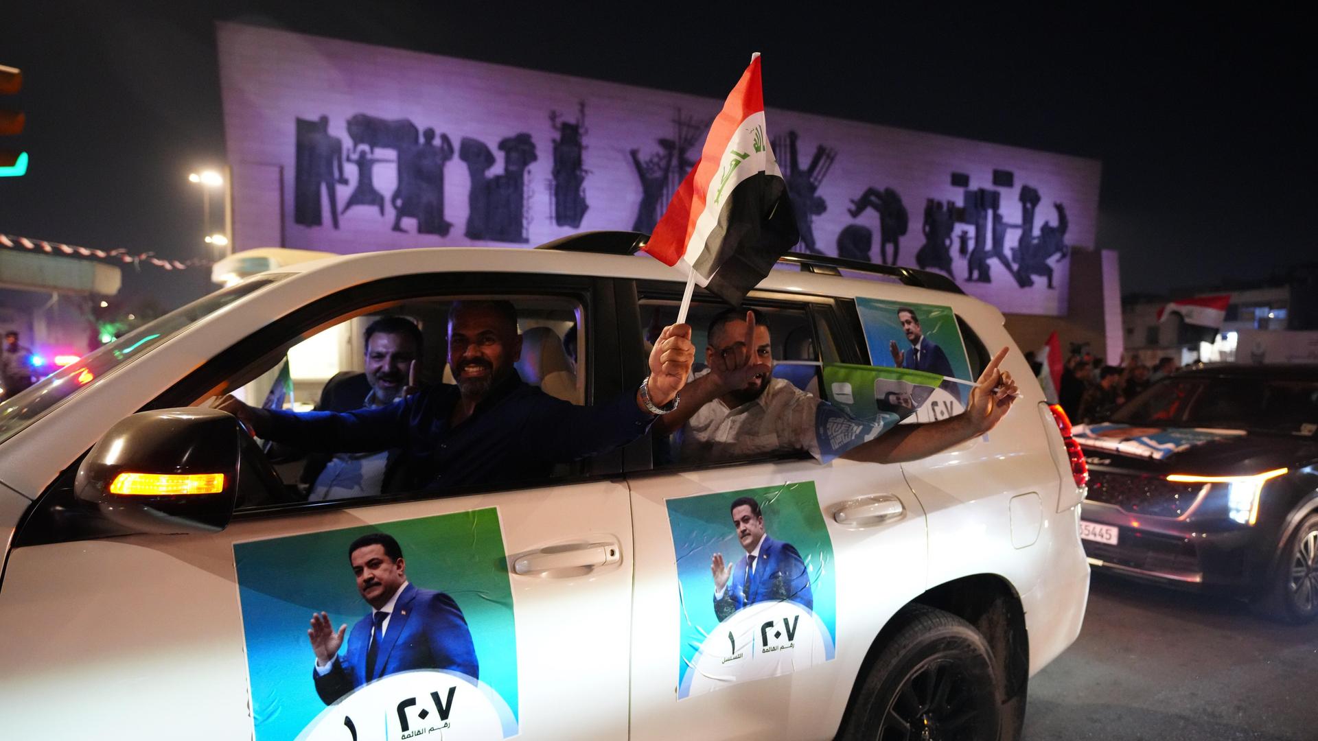 In einem weißen Auto, dass mit Fotos des irakischen Ministerpräsidenten Schia al-Sudani beklebt ist, fahren Männer durch eine nächtliche Straße, der Fahrer hält eine irakische Fahne aus dem Fenster
