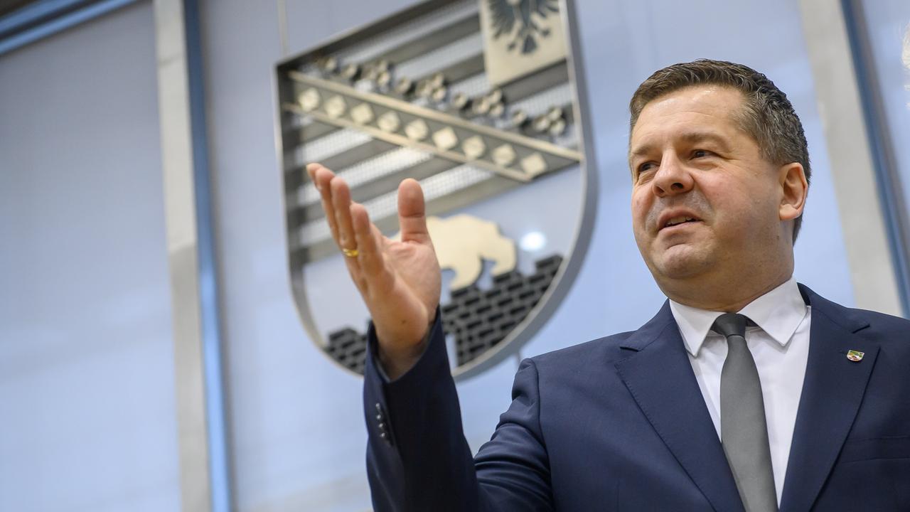 Ein Mann steht in einem großen Raum, im Hintergrund ist das Wappen des Bundeslandes Sachsen-Anhalt. Der Mann ist Sven Schulze, er wurde gerade zum Ministerpräsidenten des Landes gewählt. Er hebt seine rechte Hand nach oben und blickt zufrieden.