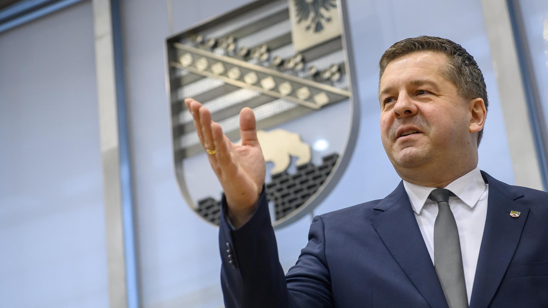 Ein Mann steht in einem großen Raum, im Hintergrund ist das Wappen des Bundeslandes Sachsen-Anhalt. Der Mann ist Sven Schulze, er wurde gerade zum Ministerpräsidenten des Landes gewählt. Er hebt seine rechte Hand nach oben und blickt zufrieden.