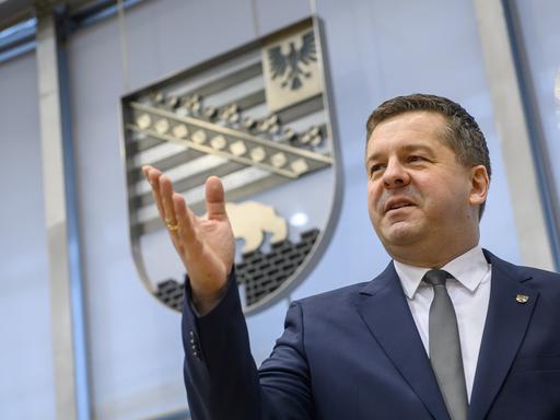 Ein Mann steht in einem großen Raum, im Hintergrund ist das Wappen des Bundeslandes Sachsen-Anhalt. Der Mann ist Sven Schulze, er wurde gerade zum Ministerpräsidenten des Landes gewählt. Er hebt seine rechte Hand nach oben und blickt zufrieden.