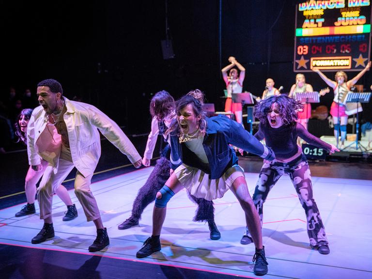 Fünf Tänzerinnen und Tänzer stehen am vorderen Rand einer quadratischen Bühne, sie tanzen und schreien. Im Hintergrund eine Band und eine Anzeigentafel für einen Wettkampf: Team "Alt" gegen Team "Jung".