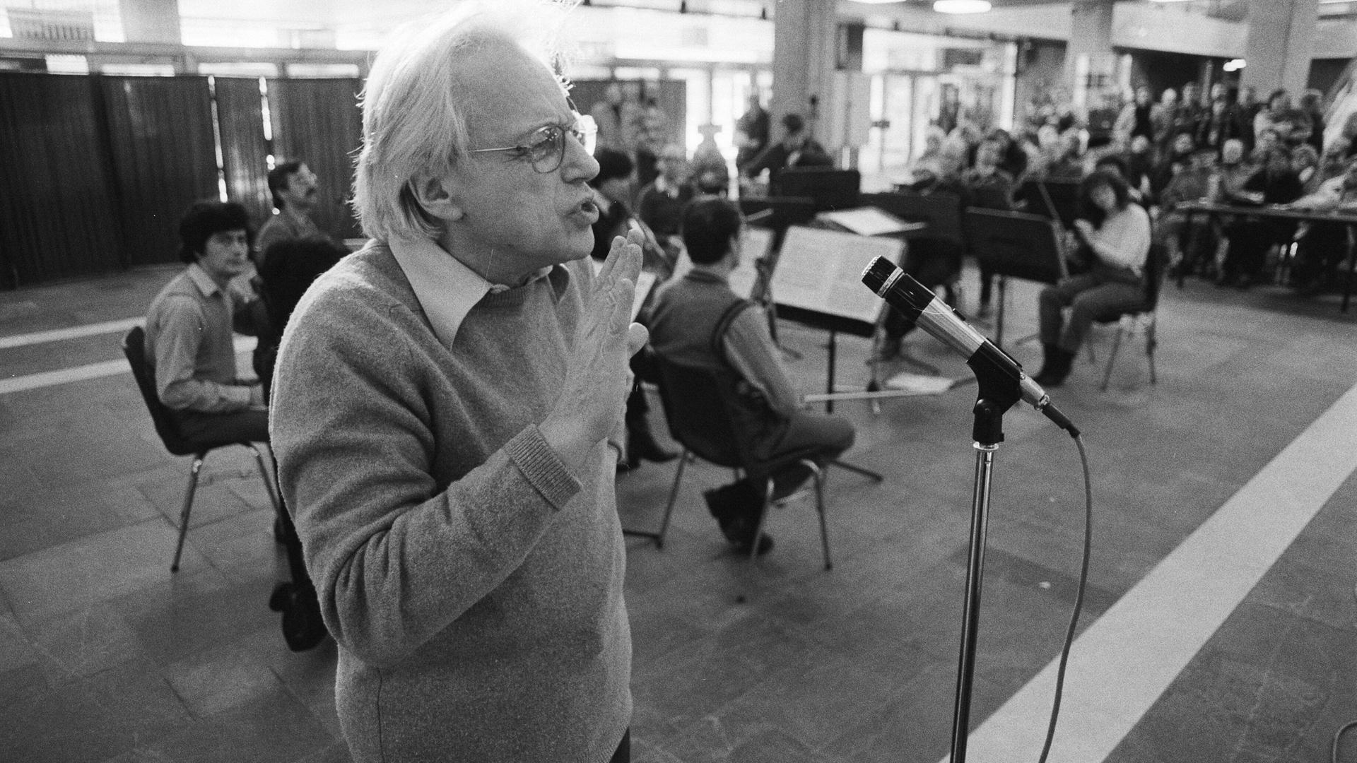 Der Komponist György Ligeti steht vor einem Mikrofon und scheint etwas zu sagen. 