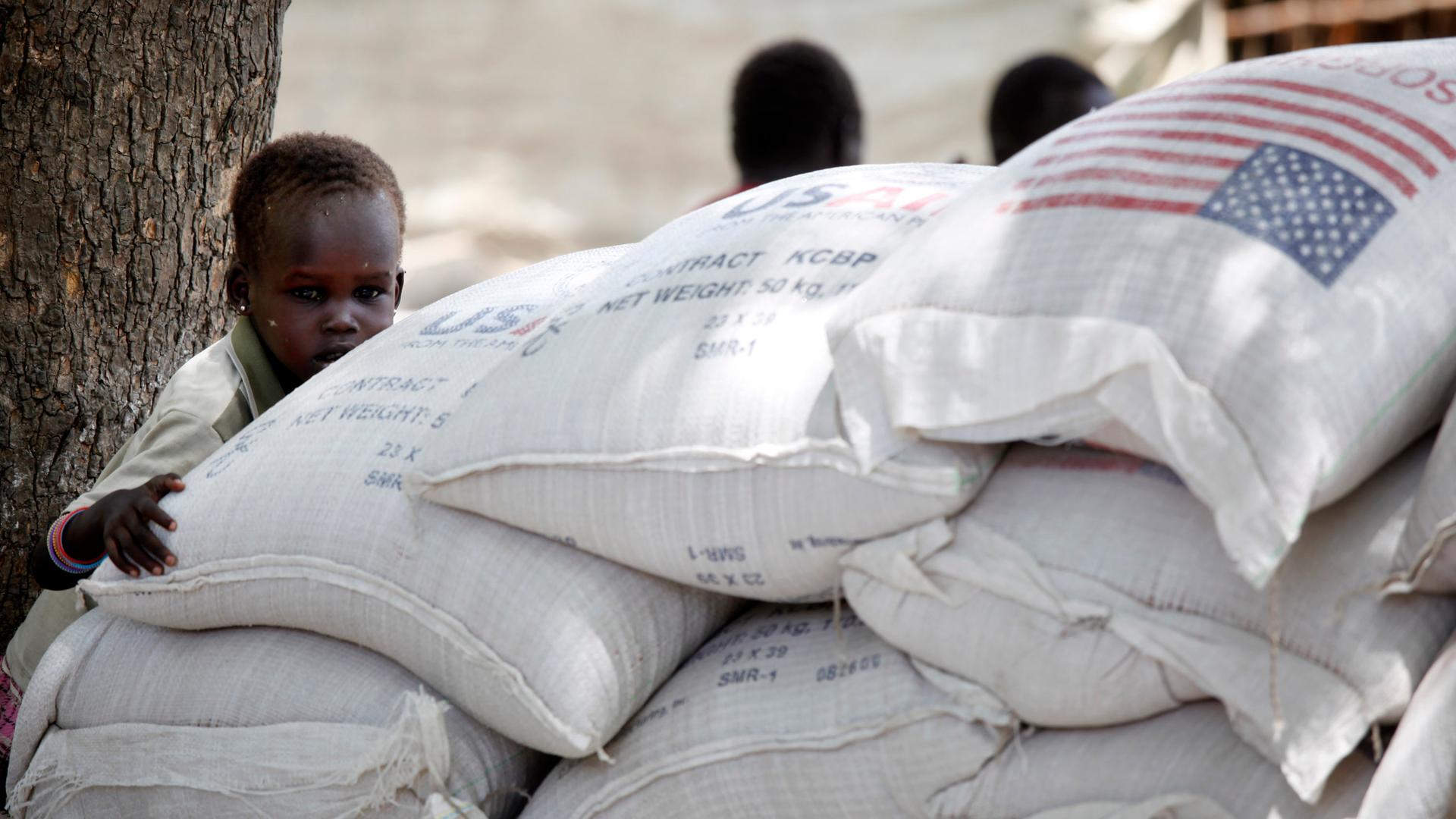 Ein kleines Kind spielt auf weißen Säcken, auf denen die Aufschrift Sorghum, das Logo von USAID sowie die US-Flagge zu erkennen sind