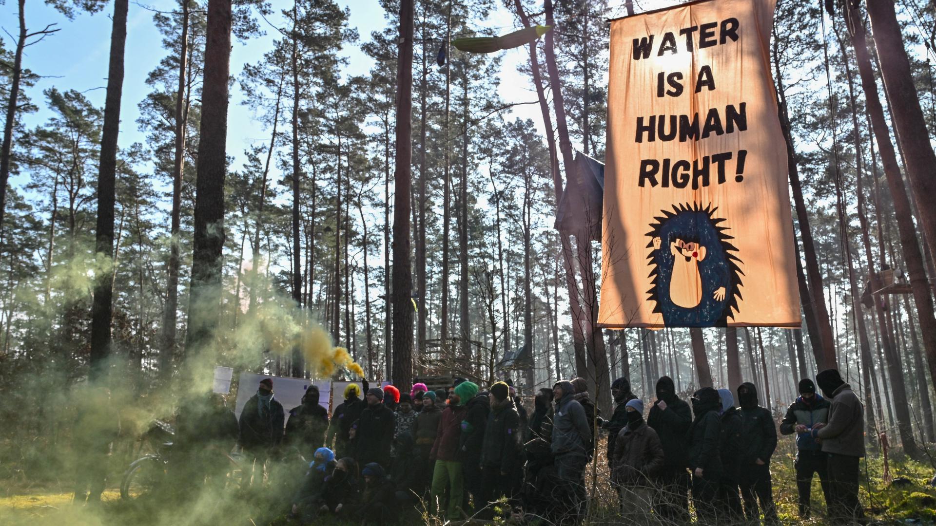 Grünheide: Aktivisten der Initiative "Tesla stoppen" stehen mit Rauchfackeln vor einem Transparent mit der Aufschrift "Water is a human right!" in einem Kiefernwald nahe dem Werk der Tesla-Gigafactory Berlin-Brandenburg.