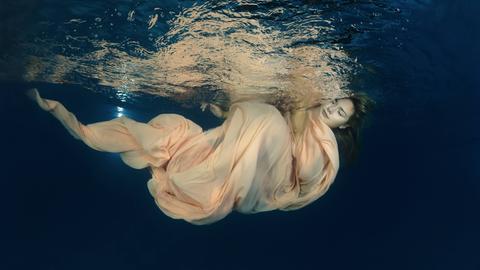 Eine Frau in einem wallenden Kleid unter der Wasseroberfläche, ihre Augen sind geschlossen