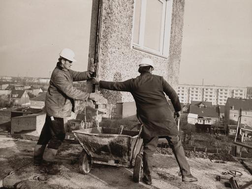 Eine Schwarz-Weiß-Aufnahme aus den 1970er Jahren zeigt Bauarbeiter beim Einfügen eines Teilstücks für ein Wohnhaus, das in Plattenbauweise errichtet wird.