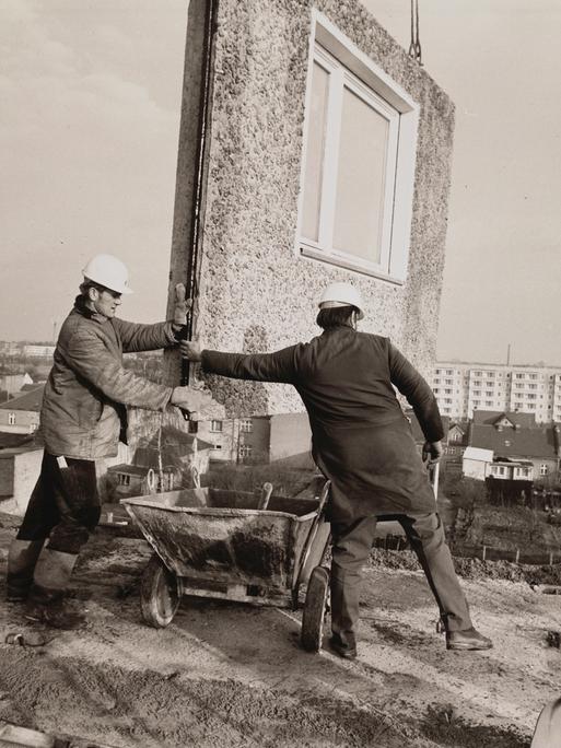 Eine Schwarz-Weiß-Aufnahme aus den 1970er Jahren zeigt Bauarbeiter beim Einfügen eines Teilstücks für ein Wohnhaus, das in Plattenbauweise errichtet wird.