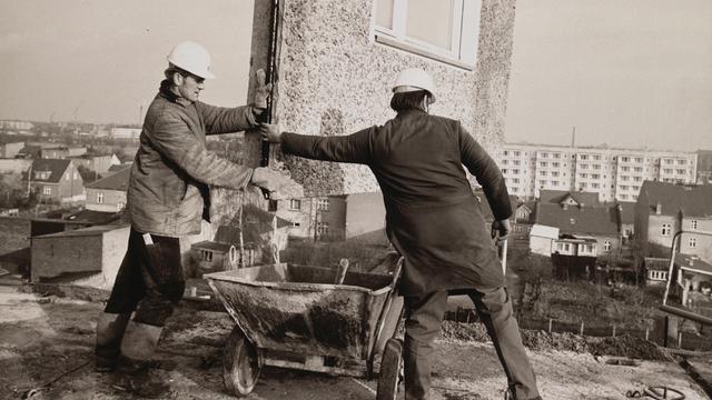 Eine Schwarz-Weiß-Aufnahme aus den 1970er Jahren zeigt Bauarbeiter beim Einfügen eines Teilstücks für ein Wohnhaus, das in Plattenbauweise errichtet wird.