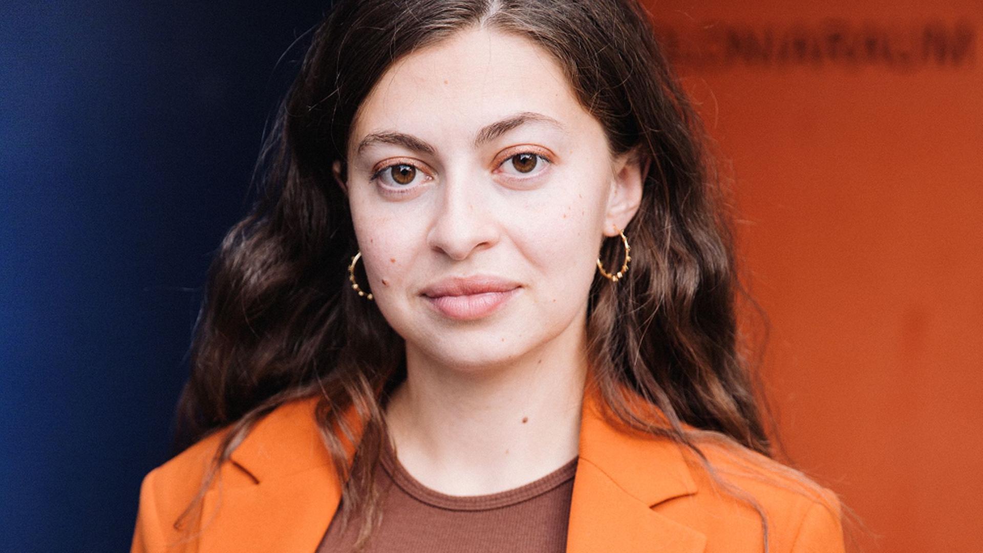 Porträt von Luna Al-Mousli. Eine junge Frau mit glatten, dunkelbraunen Haaren, die einen orangen Blazer trägt.