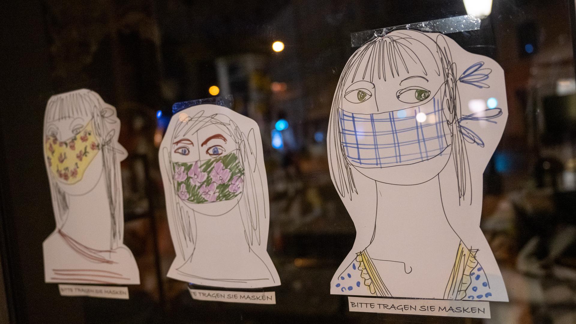 Am Schaufenster eines Geschäfts kleben drei auf Papier gezeichnete und ausgeschnittene Köpfe, darunter jeweils der handschriftliche Hinweis: "Bitte tragen Sie Masken".