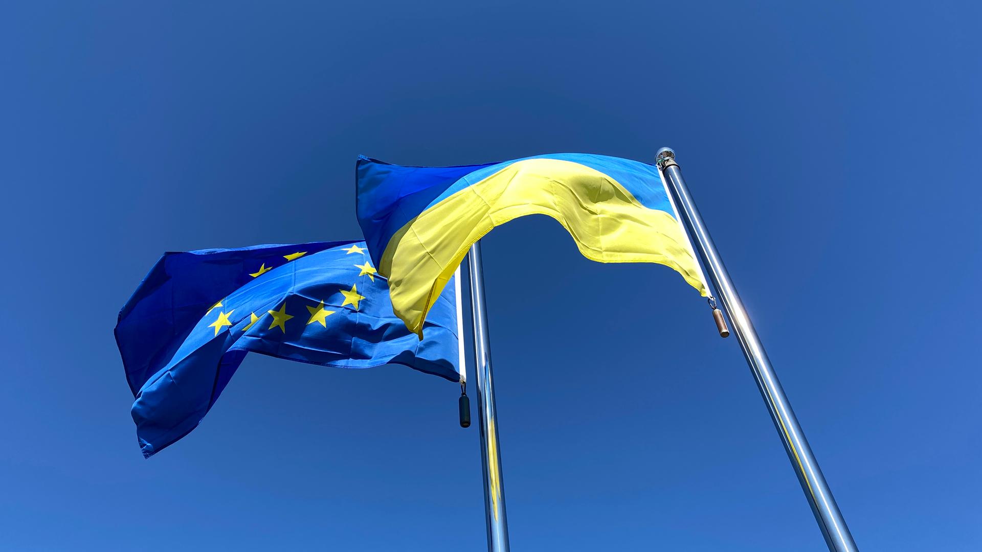 Die Flaggen der Ukraine und der Europäischen Union wehen an Fahnenmasten vor blauem Himmel.