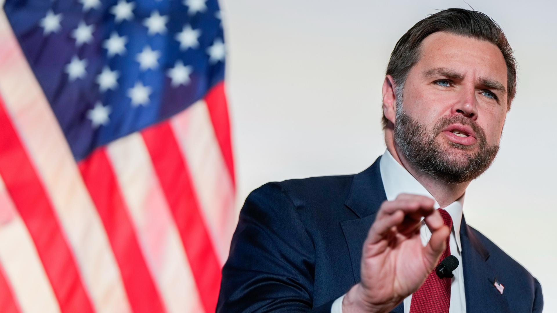 Der bärtige Vance spricht vor einer amerikanischen Flagge. Im Hintergrund eine weiße Wand. Er trägt ein schwarzes Jackett.