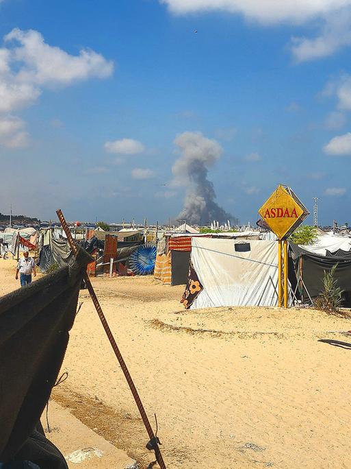 Notdürftige Zelte stehen im Sand. Ein unbefestigter Weg führt hindurch. Im Hintergrund steigt offenbar nach einer Explosion Rauch auf.