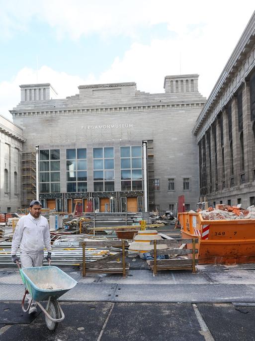 Blick auf die Baustelle des Pergamonmuseums: Ein Bauarbeiter fährt eine Schubkarre, im Hintergrund sind Gerüstteile und ein Abfallcontainer zu sehen.