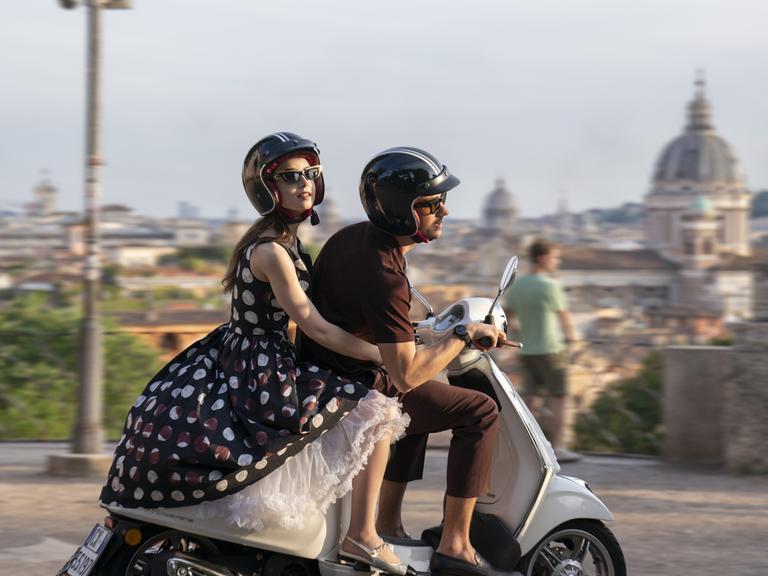 Die Schauspieler Lily Collins und Eugenio Franceschini sitzen auf einem Motorroller und fahren durch Rom. 