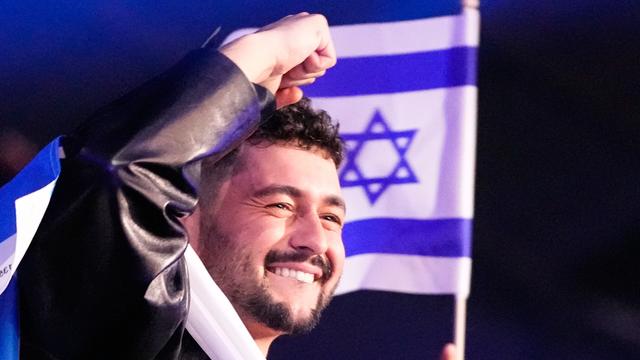 Noam Bettan macht mit seiner rechten Hand eine Faust und lächelt. Hinter ihm ist die Fahne von dem Land Israel.