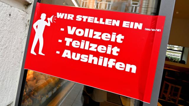 Auf einem roten Schild an der Glastür einer Bäckerei steht: "Wir stellen ein (m/w/d): Vollzeit, Teilzeit, Aushilfe"