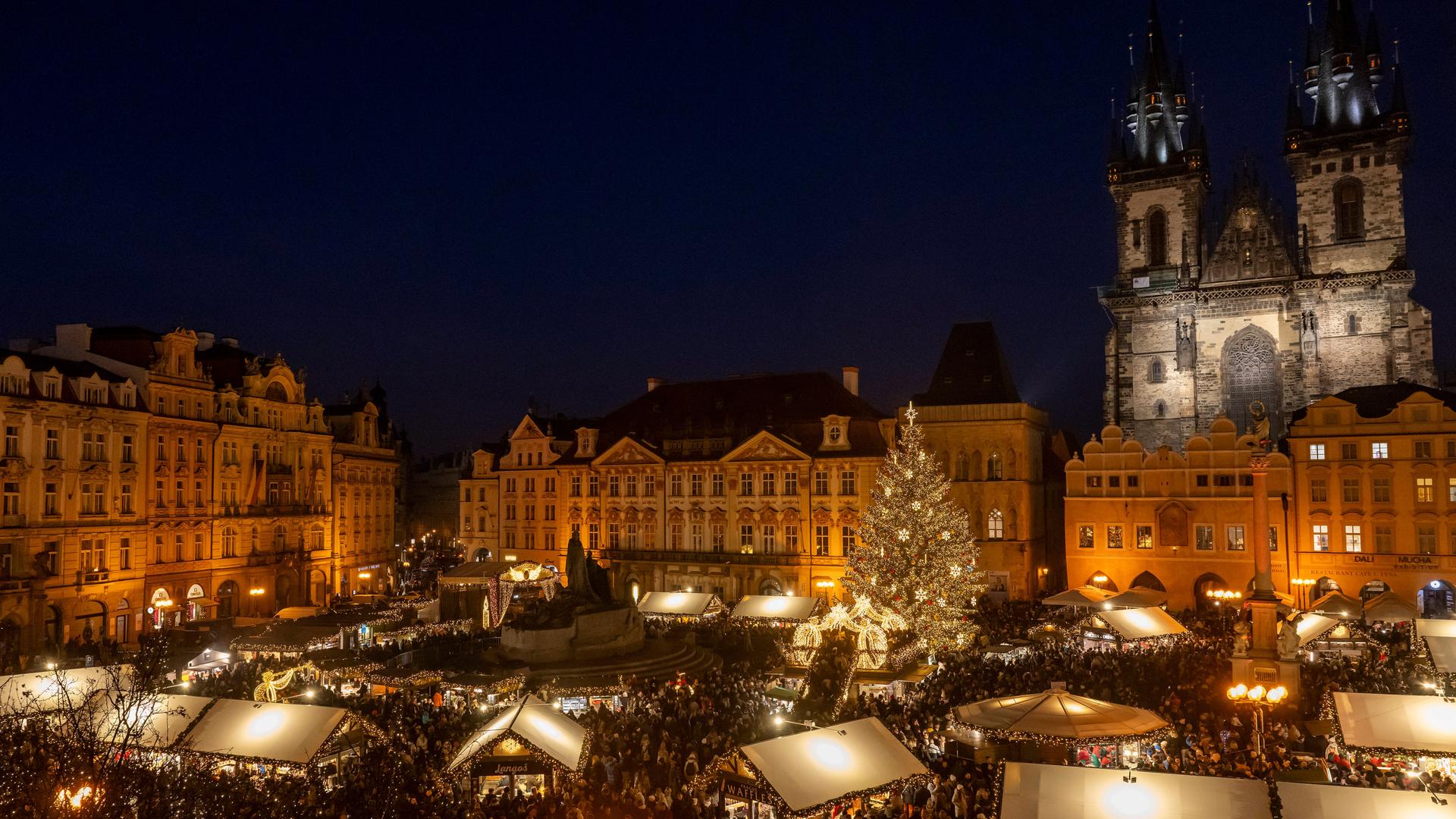 Blick von leicht oben auf einen Weihnachtsmarkt bei Nacht im Zentrum von Prag. Dahinter sieht man die angestrahlten  Bauten der historische Altstadt und rechts eine Kirche.