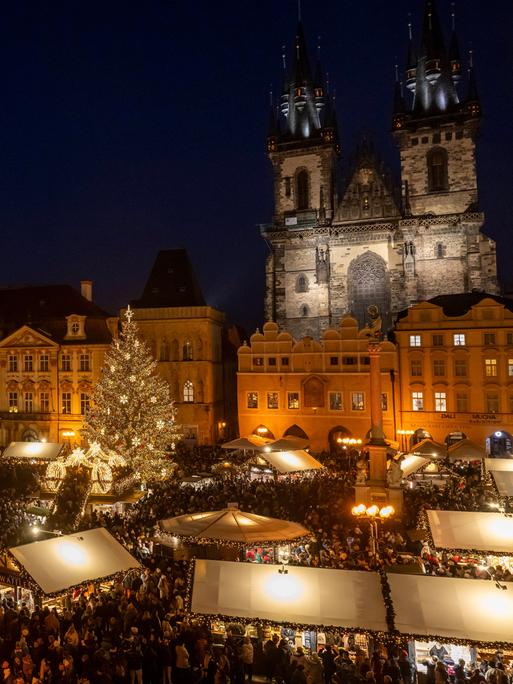 Blick von leicht oben auf einen Weihnachtsmarkt bei Nacht im Zentrum von Prag. Dahinter sieht man die angestrahlten  Bauten der historische Altstadt und rechts eine Kirche.