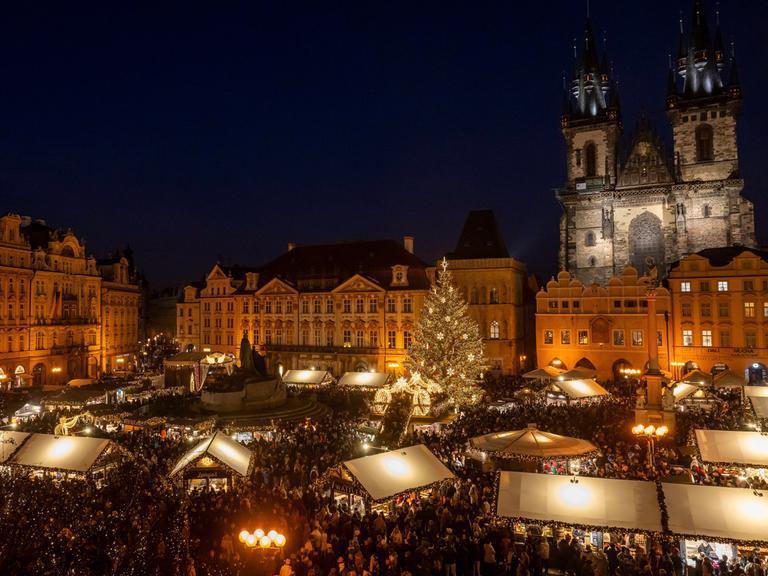 Blick von leicht oben auf einen Weihnachtsmarkt bei Nacht im Zentrum von Prag. Dahinter sieht man die angestrahlten  Bauten der historische Altstadt und rechts eine Kirche.