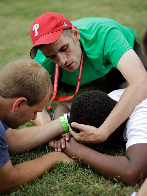 Die Betreuer halten den Kopf und die Hände eines 13-jährigen Campteilnehmers fest, um zu verhindern, dass er sich verletzt, wenn er einen Tic hat, bei dem er plötzlich zu Boden fällt und mit Kopf und Händen auf dem Gras aufschlägt. Die Betreuer halten den Kopf und die Hände eines 13-jährigen Campteilnehmers fest, um zu verhindern, dass er sich verletzt, wenn er einen Tic hat, bei dem er plötzlich zu Boden fällt und mit Kopf und Händen auf dem Gras aufschlägt.
