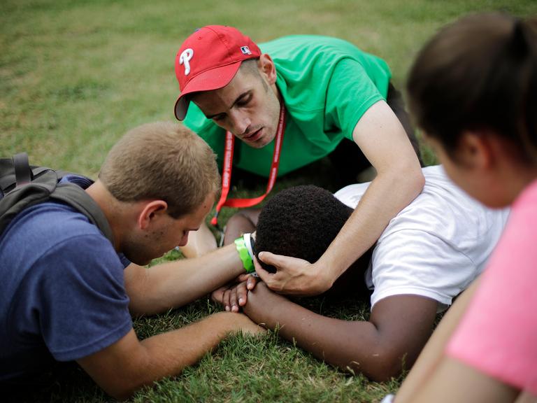 Die Betreuer halten den Kopf und die Hände eines 13-jährigen Campteilnehmers fest, um zu verhindern, dass er sich verletzt, wenn er einen Tic hat, bei dem er plötzlich zu Boden fällt und mit Kopf und Händen auf dem Gras aufschlägt. Die Betreuer halten den Kopf und die Hände eines 13-jährigen Campteilnehmers fest, um zu verhindern, dass er sich verletzt, wenn er einen Tic hat, bei dem er plötzlich zu Boden fällt und mit Kopf und Händen auf dem Gras aufschlägt.