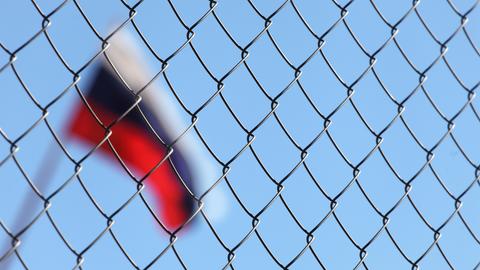 Durch einen Maschendrahtzaun hindurch ist im Hintergrund unscharf eine wehende russische Flagge zu erkennen.