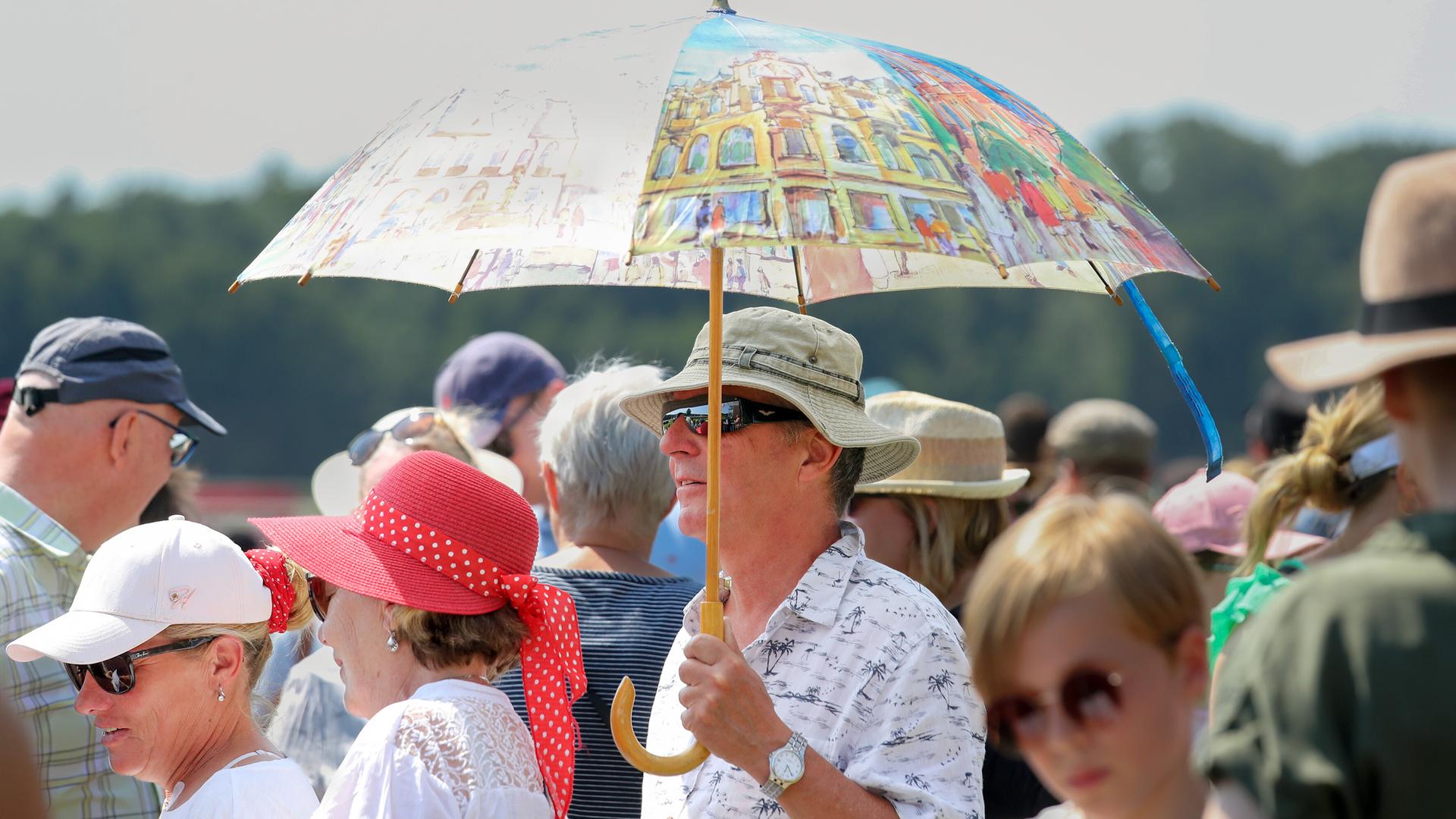 Mann schützt sich mit einem großen bunten Regenschirm gegen die Sonne, inmitten einer Menschenmenge im Berliner Hoppegarten Mann schützt sich mit einem großen bunten Regenschirm gegen die Sonne, inmitten einer Menschenmenge im Berliner Hoppegarten