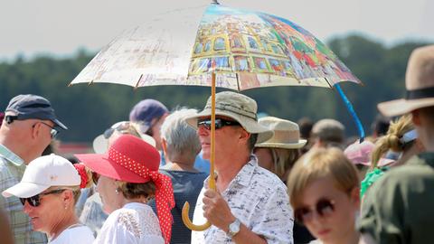 Mann schützt sich mit einem großen bunten Regenschirm gegen die Sonne, inmitten einer Menschenmenge im Berliner Hoppegarten