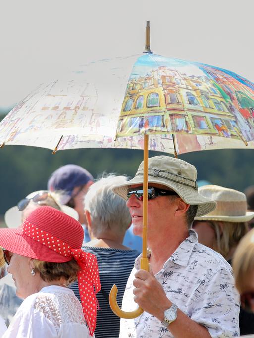 Mann schützt sich mit einem großen bunten Regenschirm gegen die Sonne, inmitten einer Menschenmenge im Berliner Hoppegarten