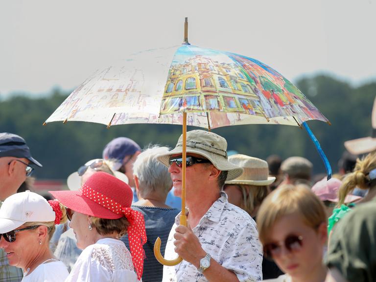 Mann schützt sich mit einem großen bunten Regenschirm gegen die Sonne, inmitten einer Menschenmenge im Berliner Hoppegarten