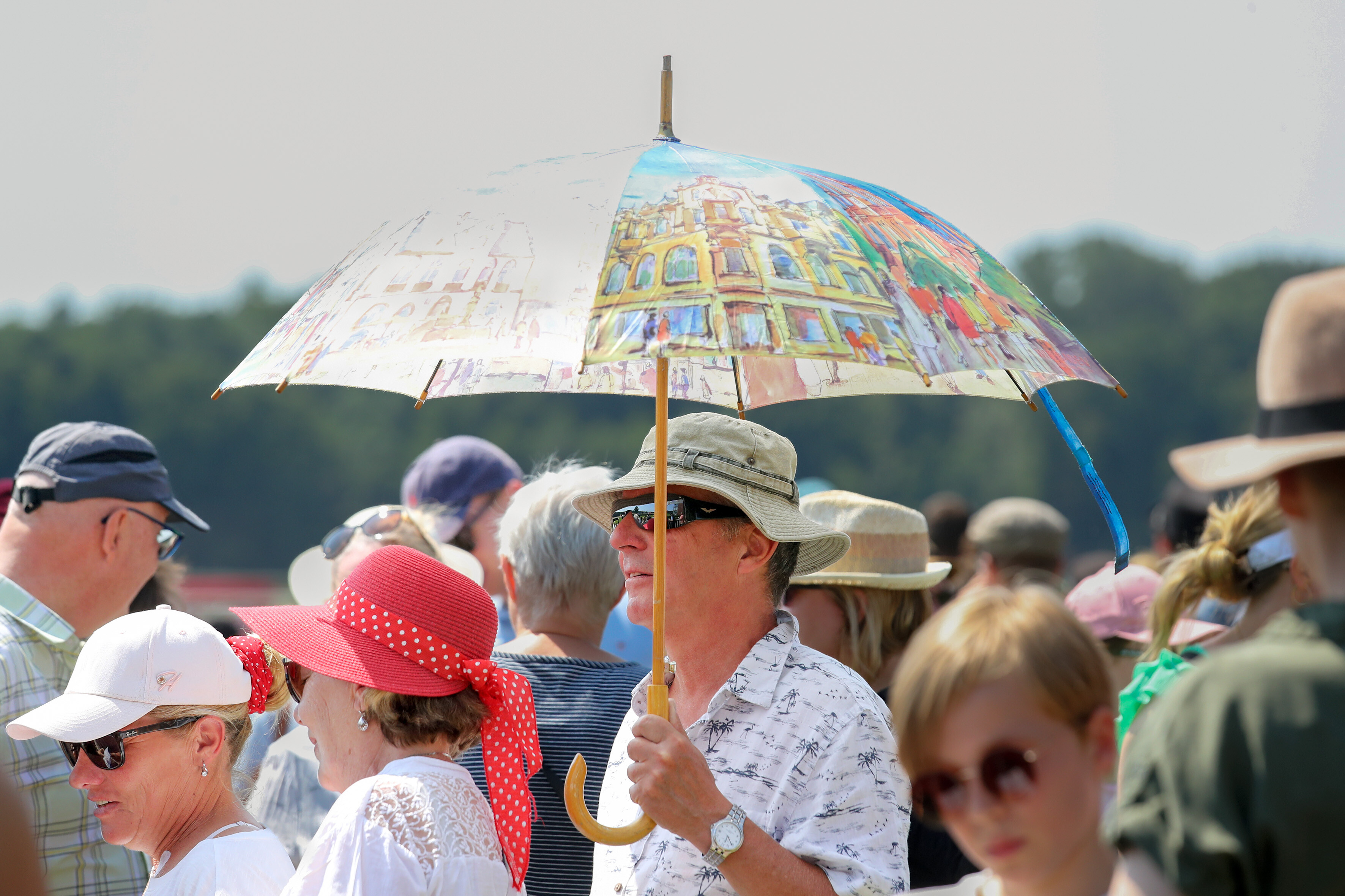 Mann schützt sich mit einem großen bunten Regenschirm gegen die Sonne, inmitten einer Menschenmenge im Berliner Hoppegarten