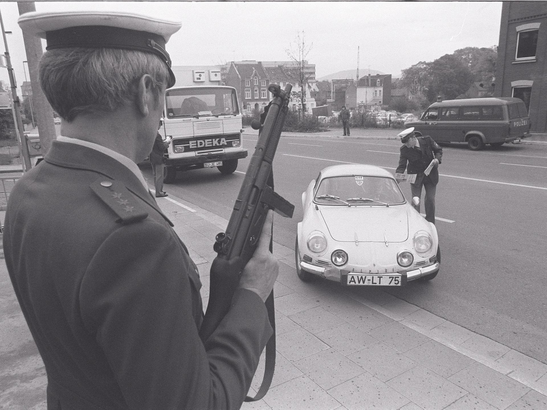 Eine Polizeikontrolle in den 70er-Jahren. Ein Polizist posiert mit einem Maschinengewehr. Ein anderer Polizist verteilt Flugblätter an die Insassen eines angehaltenen Autos.