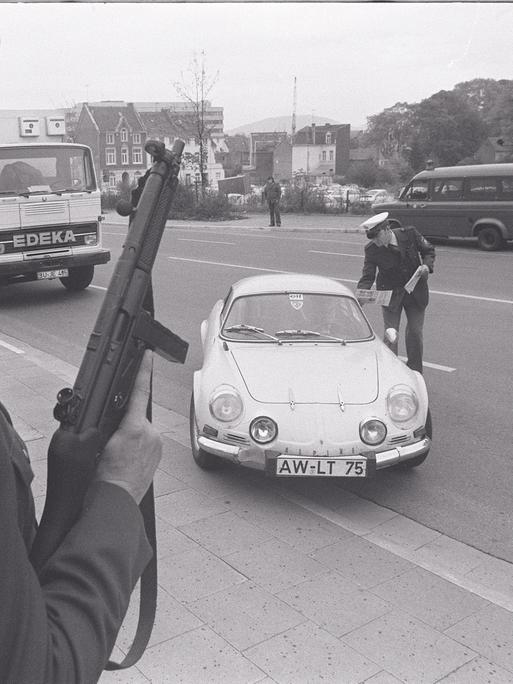 Eine Polizeikontrolle in den 70er-Jahren. Ein Polizist posiert mit einem Maschinengewehr. Ein anderer Polizist verteilt Flugblätter an die Insassen eines angehaltenen Autos.