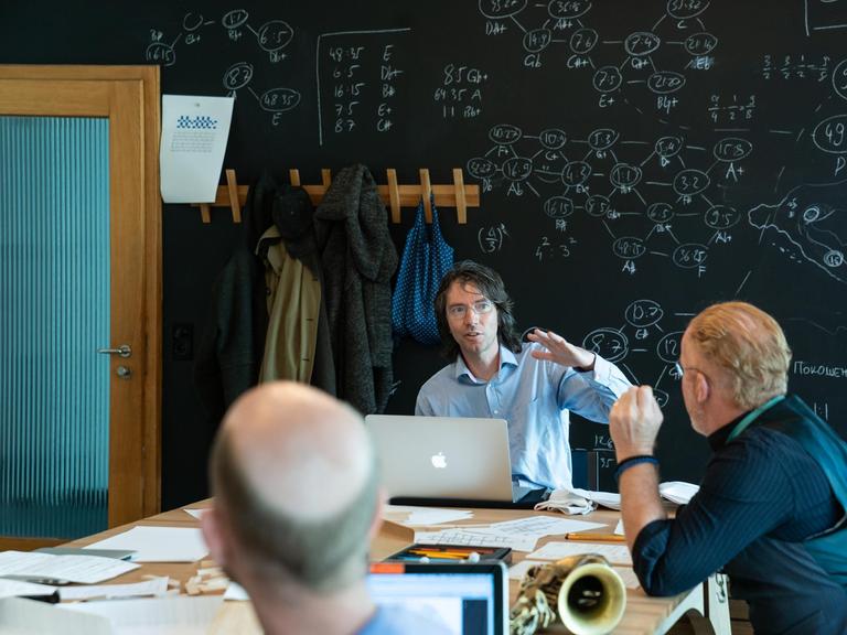 Der Komponist sitzt vor einer Tafel mit Formeln und ist mit Seminarteilnehmern im Gespräch