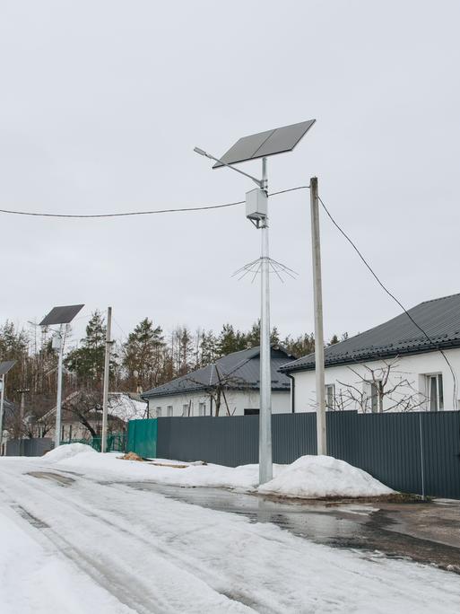 Blick in eine verschneite Straße mit Einfamilienhäusern. An den Strommasten sind Solarpaneele installiert.
