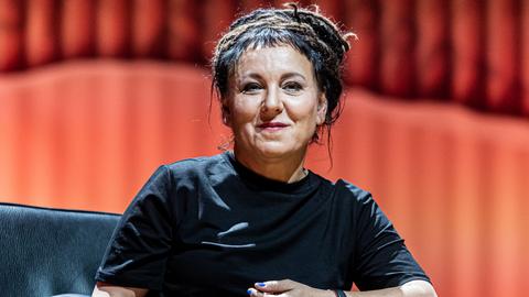 Olga Tokarczuk sitzt auf einem Stuhl auf einer Bühne und blickt lächelnd in die Kamera.